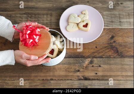 Les mains des femmes avec une boîte à biscuits en forme de coeur sur une base en bois et une assiette avec des biscuits. Concept Saint-Valentin, Fête des mères, anniversaire. Banque D'Images