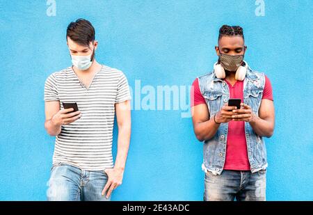 Amis masculins multiraciaux avec masques faciaux utilisant l'application de suivi avec Smartphones mobiles - jeunes gens de millénial regardant les mises à jour sur réseau social Banque D'Images