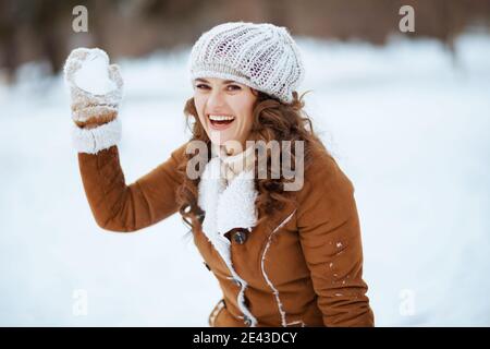 femme moderne souriante de 40 ans avec moufles dans un bonnet tricoté et un manteau en peau de mouton jouant au boules de neige à l'extérieur dans le parc de la ville en hiver. Banque D'Images