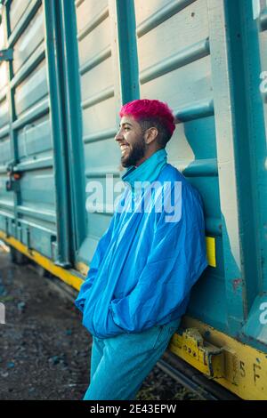 Beau homme aux cheveux teints de rose, portant une veste rétro s'inclinant contre un wagon de chemin de fer Banque D'Images