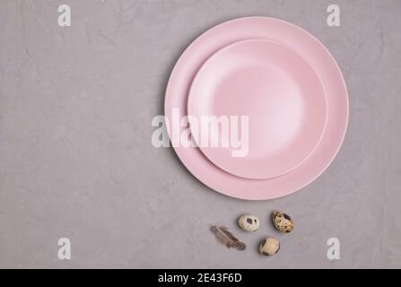 Table à poser à plat en gros plan avec deux assiettes vides roses et de petits œufs de caille avec une plume. Fond en béton gris. Concept de Pâques. Séle Banque D'Images