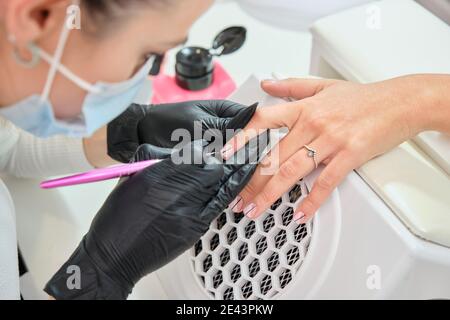 Gros plan de la récolte maître anonyme en gants avec cuticule métallique pousseur tout en faisant la manucure pour femme client dans le salon de beauté Banque D'Images