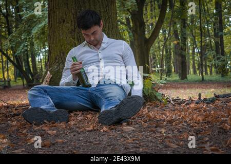 L'homme ivre déprimé se trouve sur le sol sous l'arbre avec une bouteille de bière Banque D'Images