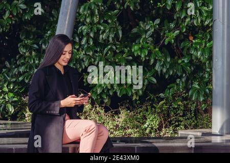 Ensemble complet de jeune femme souriante dans une tenue élégante assis sur la terrasse et la tablette tout en vérifiant les informations pour projet d'entreprise Banque D'Images