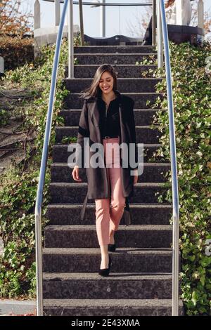 Corps entier petit angle de jeune femme moderne souriante manteau noir tendance au rez-de-chaussée dans le parc de la ville Banque D'Images