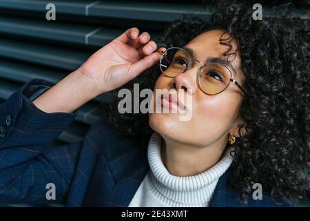 Jeune femme ethnique sérieuse auto-assurée dans des vêtements élégants se touchant lunettes et regarder vers le haut tout en se tenant contre le mur rayé gris Banque D'Images