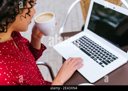Par le dessus, une femme d'affaires ethnique non reconnaissable, assise à table pour boire un café tout en travaillant à distance sur un ordinateur portable Banque D'Images