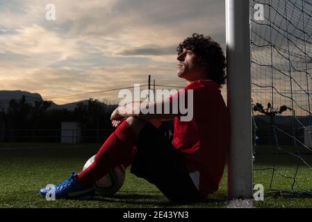 Vue latérale complète du joueur de football pensif avec ballon assis près de l'objectif sur le terrain de sport et en regardant loin soirée Banque D'Images