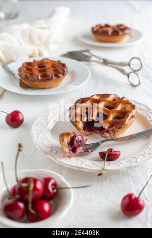 Vue latérale de trois plaques blanches de cerises individuelles avec une tarte coupée ouverte avec une fourchette, un fond blanc et des cerises et un ustensile de service argenté i Banque D'Images