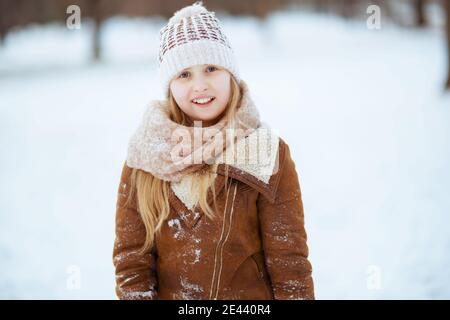 Portrait d'un enfant heureux et élégant en plein air dans le parc de la ville en hiver dans un bonnet tricoté et un manteau de peau de mouton jouant. Banque D'Images