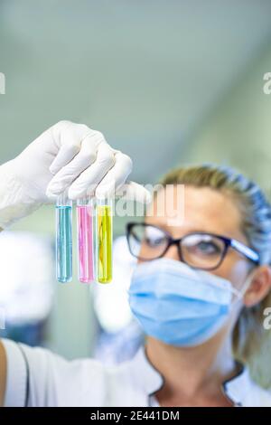 Femme scientifique sérieuse dans le masque et les gants de latex debout avec tubes à essai en verre remplis de liquides chimiques de différentes couleurs et de travailler en laboratoire Banque D'Images