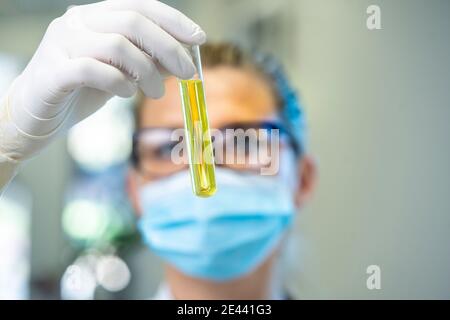 Femme scientifique sérieuse dans le masque et les gants de latex debout avec tube à essai en verre rempli de liquide chimique pendant le travail laboratoire Banque D'Images