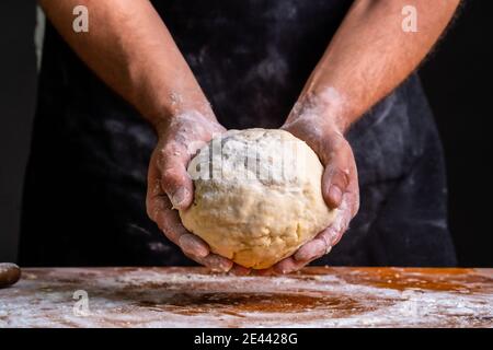 Rognez un boulanger mâle méconnu qui tient la pâte dans les mains en étant debout à la table en bois dans la boulangerie Banque D'Images