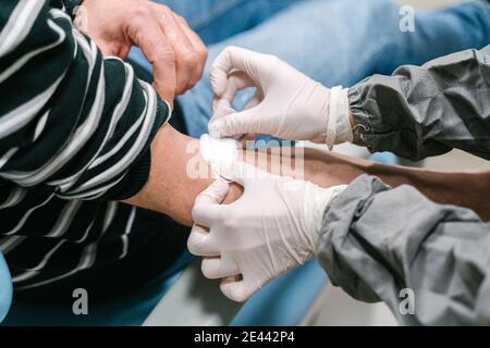 Vue latérale grand angle de la récolte anonyme infirmière en latex gants appliquant le patch sur le bras du patient après l'exécution du sang test en clinique Banque D'Images