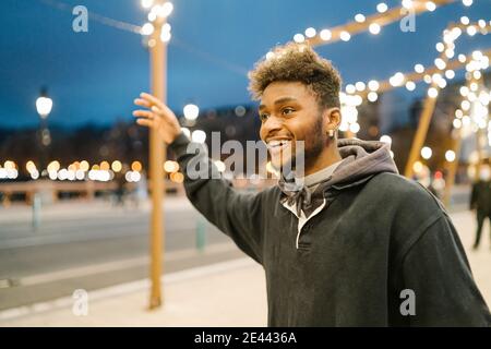 Gai jeune barbu afro-américain hippster homme voyageur avec bras élevé prenant un taxi dans la rue urbaine le soir Banque D'Images