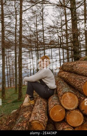 Jeune femme souriante dans des vêtements chauds et chapeau regardant appareil photo assis sur un tas de bois empilés les uns sur les autres en campagne Banque D'Images