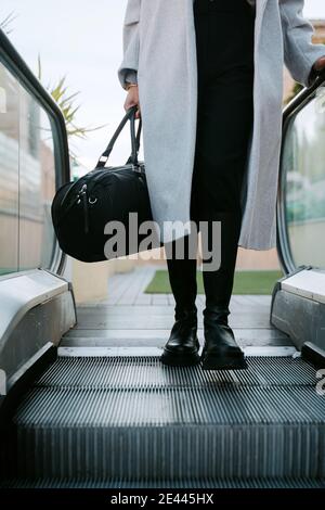 Femme d'affaires élégante et sans visage sous un manteau et avec sac sur l'escalier roulant dans le centre-ville Banque D'Images