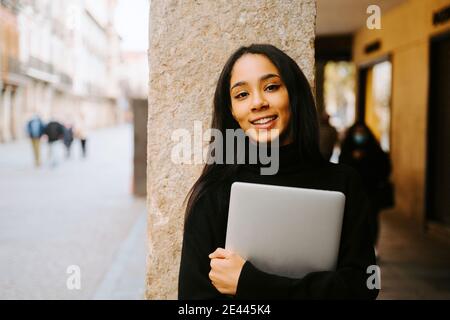 Charmante jeune femme ethnique avec netbook souriant à l'appareil photo colonne de pierre sur la rue de la ville Banque D'Images