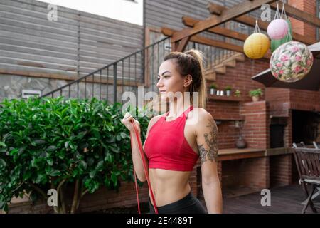 Une athlète féminine gaie dans des vêtements de sport faisant de l'exercice de boucle de biceps avec bande élastique pendant l'entraînement dans la cour et vue sur l'extérieur Banque D'Images
