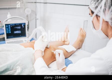 Attention sélective sur les mains d'un médecin portant un masque, un capuchon, des gants et un uniforme effectuant un test d'échographie sur les jambes d'un patient allongé sur une civière Banque D'Images