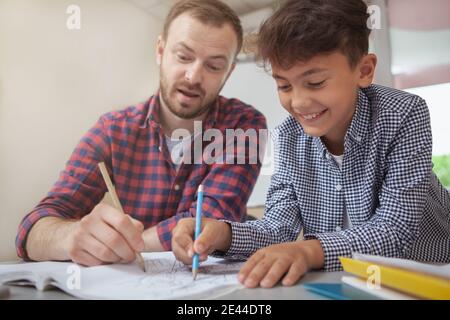 Plan court d'un charmant garçon d'école souriant joyeusement, en train de dessiner avec son professeur pendant la classe d'art. Enseignant aidant son petit étudiant avec l'artiste Banque D'Images