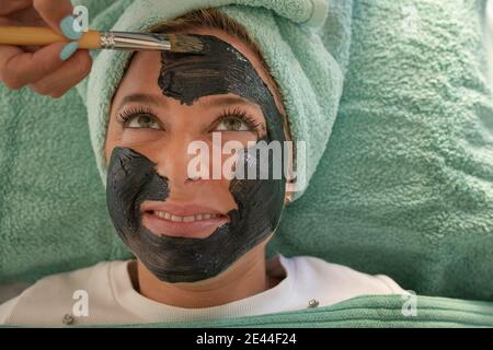 Du dessus de la récolte femelle anonyme cosmetologiste appliquant de l'argile noire masque sur le visage d'une femme souriante et heureuse couché sur un canapé avec une serviette sur la tête pendant la peau Banque D'Images