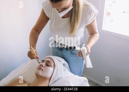 De dessus de la récolte jeune femme cosméticienne dans des vêtements décontractés appliquant un masque blanc sur le visage de la femme allongée détendue pendant la procédure de spa dans le salon Banque D'Images