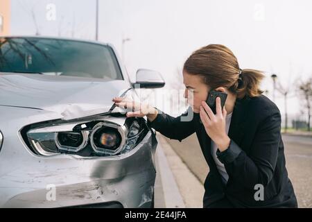 Contrarié jeune femme pilote dans une tenue décontractée ayant conversation téléphonique sur route et inspection des dommages sur la voiture moderne après collision Banque D'Images