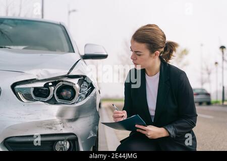 Vue latérale d'une femme agent d'assurance dans un article de tenue classique protocole à proximité d'une voiture endommagée garée dans la rue après l'accident Banque D'Images