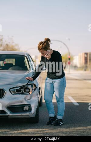 Ensemble complet de jeune femme pilote contrarié dans une tenue décontractée avoir une conversation téléphonique en étant debout sur la route et en inspectant les dommages sur la voiture moderne après cr Banque D'Images