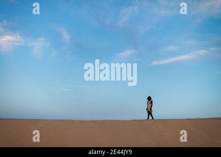 Vue latérale d'une femme africaine anonyme en robe légère marchant sur les sables du désert Banque D'Images