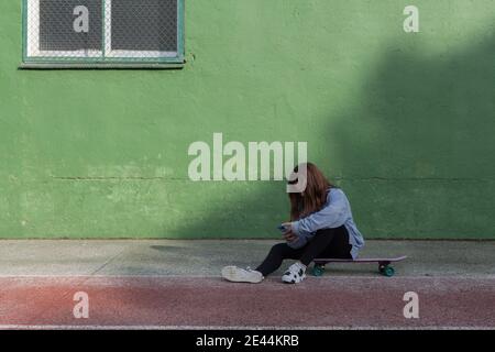 Corps complet de femme adolescente fatiguée dans une tenue décontractée assis sur le panneau de penny près du mur vert et de la navigation de téléphone mobile après l'entraînement en plein air Banque D'Images