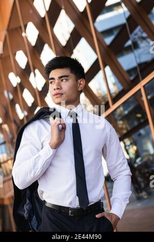 Homme d'affaires asiatique sérieux et bien habillé debout avec veste dans le centre d'affaires et en regardant loin Banque D'Images