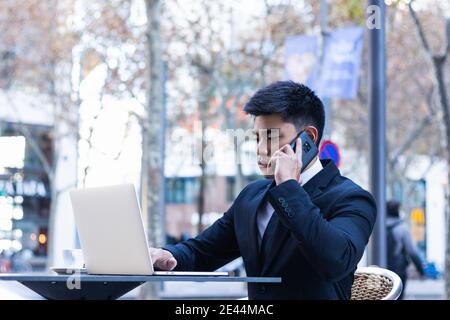 Homme d'affaires asiatique souriant assis à table dans le café de rue et travailler à distance en tapant sur un ordinateur portable et en parlant smartphone Banque D'Images