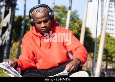 Jeune homme ethnique concentré dans un élégant rouge à capuche à l'écoute musique dans un casque sans fil et lecture de journaux intéressants tout en vous refroidissant sur gre Banque D'Images