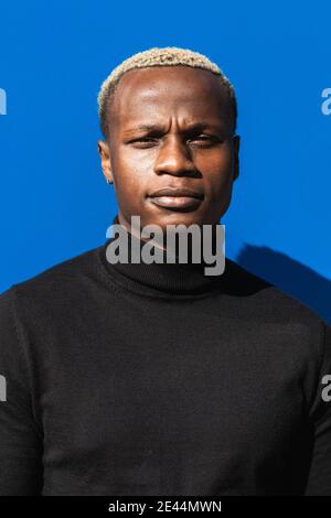 Calme jeune Africain américain avec des cheveux blonds en noir col roulé debout sur fond bleu vif le jour ensoleillé et regarder la caméra pensive Banque D'Images