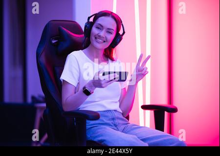 Une gamer fille en t-shirt blanc et jeans bleus tient le smartphone entre les mains et joue à l'application de jeu sur le téléphone mobile, souriant et regardant l'écran du téléphone portable Banque D'Images