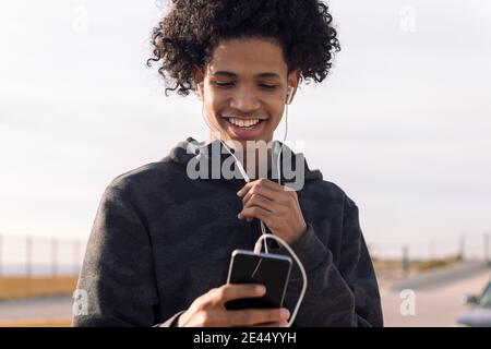 Joyeux adolescent afro-américain avec des cheveux bouclés et des écouteurs communication par téléphone mobile en vous tenant à l'opposé d'une rue floue Banque D'Images