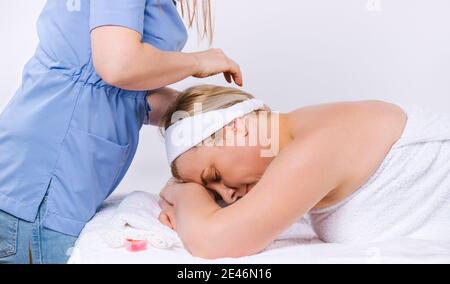 Photo rognée d'une jeune femme masseuse faisant un massage de la tête à une femme de race blanche en salon avec bougies parfumées et fond blanc. Banque D'Images