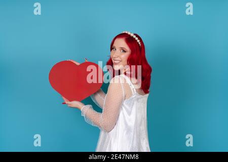 une femme joyeuse et heureuse se tient sur le côté dans une robe blanche et avec des cheveux rouges tenant un grand coeur en papier rouge isolé sur fond bleu Banque D'Images