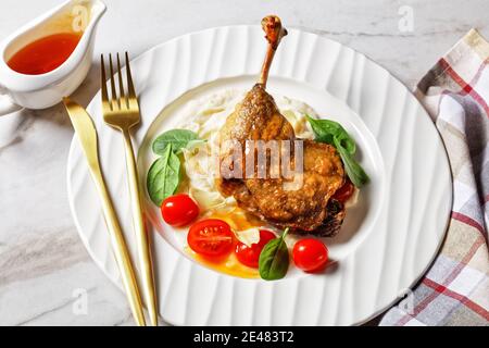 Cuisse de canard rôtie - confit de canard avec purée de panais et sauce à l'orange, tomates cerises, feuilles d'épinards fraîches servies sur une assiette blanche avec couverts dorés o Banque D'Images