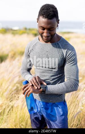 Portrait de l'homme afro-américain en bonne forme dans les vêtements de sport à l'aide de la montre intelligente tenir l'eau dans l'herbe haute Banque D'Images