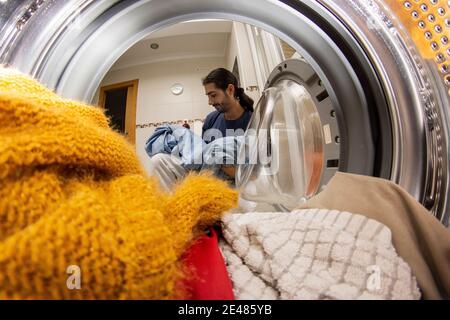 Jeune homme latin avec des cheveux longs noirs prenant les vêtements hors du lave-linge à la maison Banque D'Images