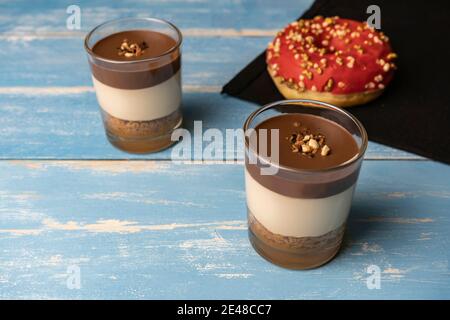 Deux desserts au chocolat en verre (yaourt ou crème, biscuits et noix) avec beignet rouge, sur fond de bois bleu. Banque D'Images