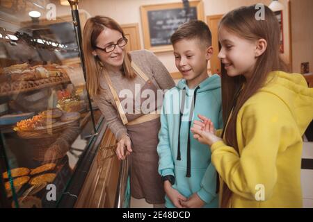 Une boulangère professionnelle gaie en tablier pour aider ses jeunes clients à choisir pâtisserie en exposition Banque D'Images