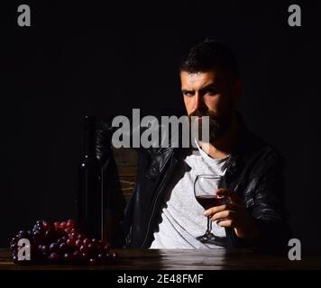 Un homme avec une barbe tient un verre de vin sur fond marron foncé. Le sommelier goûte des boissons chères. Concept de dégustation de vins et de dégustation. Le Degustator au visage grincheux se trouve à côté d'une bouteille de vin et de raisins foncés. Banque D'Images