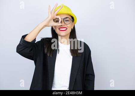 Jeune architecte femme portant un casque robuste faisant geste ok choqué avec visage souriant, oeil regardant à travers les doigts Banque D'Images