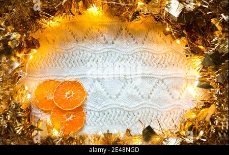 Cadre festif en guirlande dorée avec lumières et guirlande, tranches d'oranges séchées sur un tricoté blanc avec un fond de motif, espace de rédaction. Nouvelle année, Banque D'Images