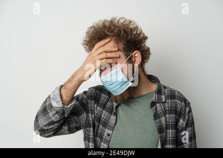 Souffrir de maux de tête portant un masque médical stérile de protection sur son visage pour protéger le coronavirus cheveux bouclés jeune homme portant du tissu écossais chemise et olive Banque D'Images