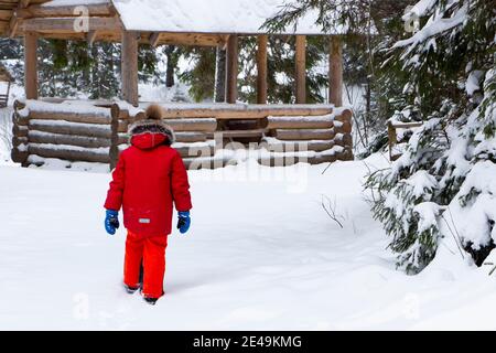 Un enfant dans une veste rouge marche dans la forêt enneigée près des arbres de Noël et d'une maison en bois. Banque D'Images